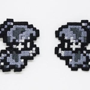 May include: Two grey and black embroidered iron-on patches featuring a pixelated character with a skull-like face.