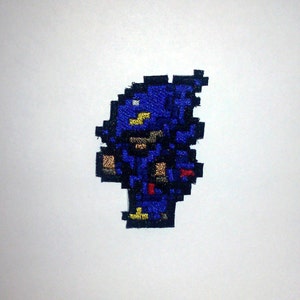 May include: A pixelated patch depicting a blue figure with yellow, brown, and red details. The design is reminiscent of a character from a classic video game, with a retro style. The patch is probably made of embroidered fabric.