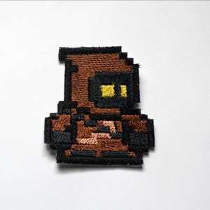 May include: A brown and black embroidered patch of a pixelated character wearing a brown hooded robe and yellow eyes.