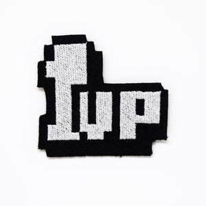 May include: A black and white embroidered patch featuring the text "1UP" in a pixelated font. The patch has a black border and is set against a white background. This patch is perfect for gamers and retro enthusiasts.