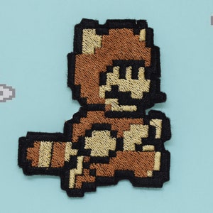 Könnte beinhalten: Ein gestickter Aufnäher aus braunem und schwarzem Stoff, der einen Videospielcharakter zeigt, der ein braunes Waschbärkostüm trägt. Der Charakter rennt nach rechts und hat einen braunen Schwanz.