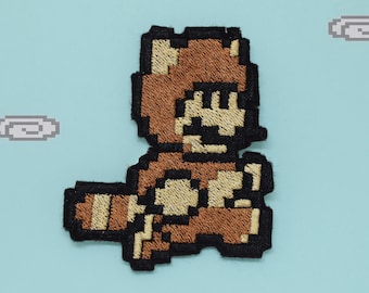 Puede incluir: Un parche bordado marrón y negro de un personaje de videojuego que lleva un disfraz de mapache marrón. El personaje corre hacia la derecha y tiene una cola marrón.