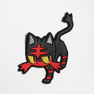 Op de afbeelding: Gestikt patch van een zwart en rood kat-achtig Pokemon-personage met gele ogen. Het personage heeft een rood kruis op zijn voorhoofd en bevindt zich in een hurkende houding.