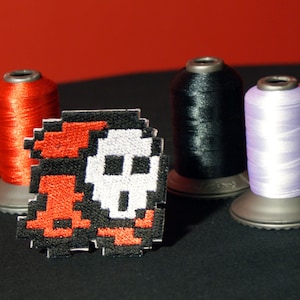 Könnte beinhalten: Ein schwarzer und weißer pixelartiger Totenkopf- und Knochen-Patch mit roten Akzenten. Der Patch liegt auf einer schwarzen Oberfläche neben drei Garnrollen: rot, schwarz und lavendel.
