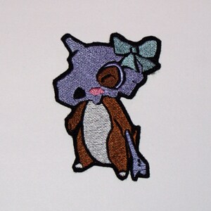 May include: Purple and brown embroidered iron-on patch of a cartoon character with a bow. The character has a skull-like head and a brown body.