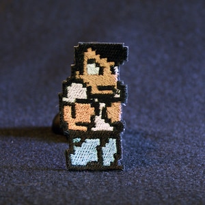 May include: A pixelated embroidered patch of a person with brown hair, a white shirt, and blue jeans. The person is looking to the left.