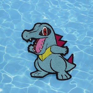 Puede incluir: Un parche bordado de un dinosaurio de dibujos animados azul y verde con una boca roja y rosa y un collar amarillo. El dinosaurio está de pie sobre un fondo azul.