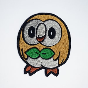 May include: Embroidered patch of a brown and white owl with green leaves around its neck. The owl has large black eyes and a small orange beak.