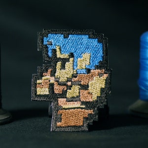 May include: A blue and brown embroidered patch featuring a pixelated character with blue hair and a brown body. The patch is made of fabric and has a black border.