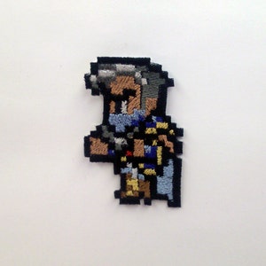 May include: Embroidered patch featuring a pixelated character from a video game. The character is a man with grey hair, a blue shirt, and a sword. The patch is made of felt and has a black border.