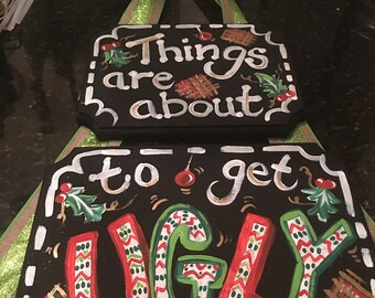 Può includere: Un cartello nero in ardesia con testo bianco che recita "Things are about to get UGLY". Il cartello è decorato con agrifoglio verde, bacche rosse e un motivo a quadri. Il cartello è attaccato a un nastro verde e oro.