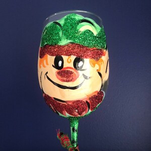 May include: A green, red, and white painted wine glass with a whimsical elf face. The elf has a green hat, red beard, and a big smile.
