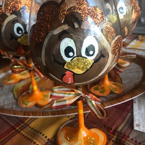 May include: Set of four wine glasses decorated with painted turkey faces. The glasses have orange and gold glitter accents and a brown, white, and orange turkey face painted on each glass. The glasses have orange stems and bases.