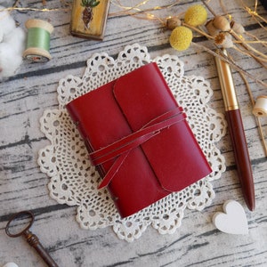 May include: A red leather journal with a brown leather strap closure, resting on a white doily. The journal is on a wooden surface with other decorative items.