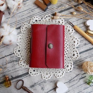 May include: A red leather journal with a flap closure and a metal button. The journal is sitting on a white doily on a wooden surface.