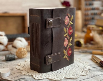 May include: A dark brown leather journal with a floral cross-stitch design in red, pink, and yellow. The journal has a flap closure with two snap buttons and a textured spine. The journal is sitting on a white lace doily.