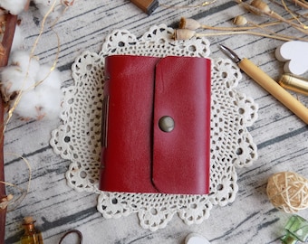 May include: A red leather journal with a flap closure and a metal button. The journal is sitting on a white doily on a wooden surface.