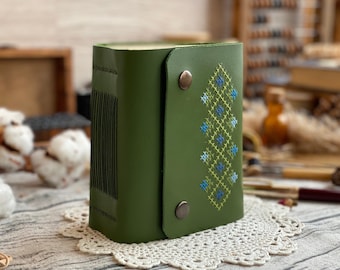 May include: A green leather journal with a flap closure and a stitched design in blue and yellow. The journal is closed with two bronze-colored snaps. The spine of the journal has vertical lines.
