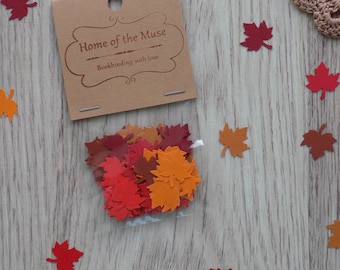 May include: A small brown paper bag with a white label that reads "Home of the Muse Bookbinding with love". The bag contains a variety of colourful paper leaves in red, orange, and brown.