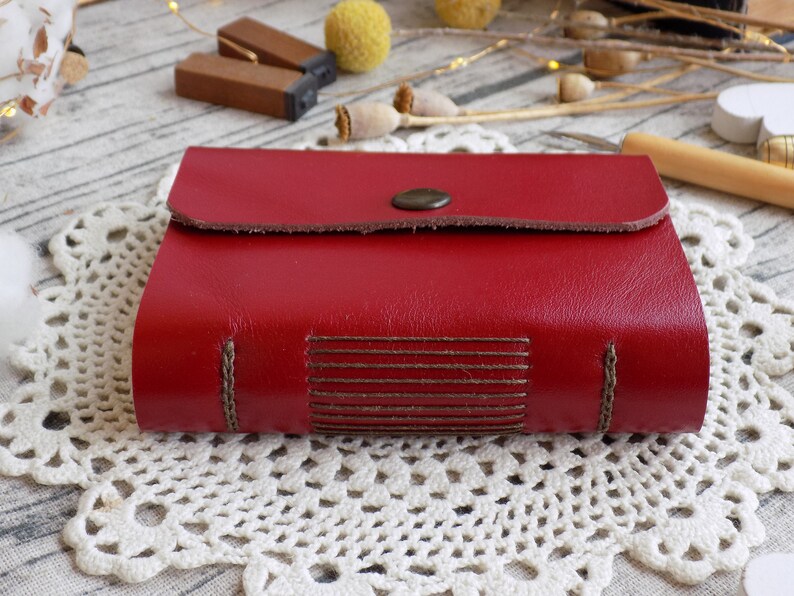 May include: A red leather-bound journal with a flap closure and a decorative button. The journal has brown stitching and a textured leather cover.