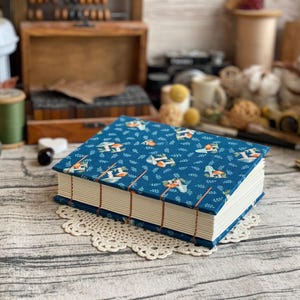 May include: A rectangular book with a blue fabric cover featuring a repeating pattern of small mushrooms and green leaves. The book has exposed stitching along the spine with visible brown thread and thick, cream-colored pages stacked neatly.