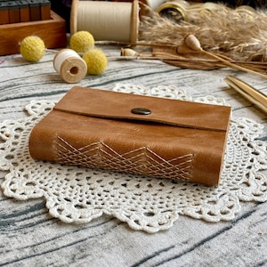 May include: A brown leather journal with a geometric stitch design and a metal button closure. The journal is resting on a white crocheted doily.