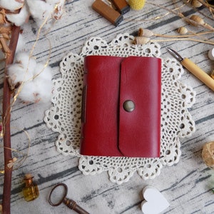 May include: A red leather journal with a flap closure and a metal button. The journal is sitting on a white doily.