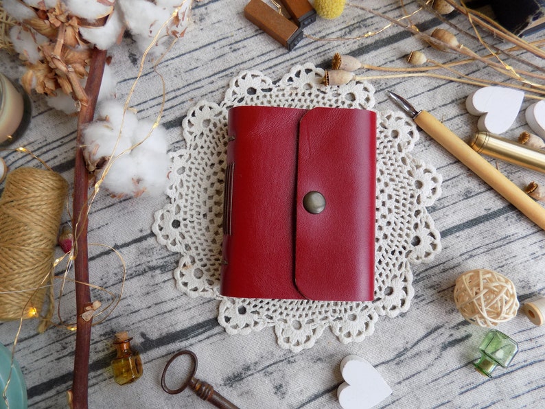 May include: A red leather journal with a flap closure and a metal button. The journal is sitting on a white doily.