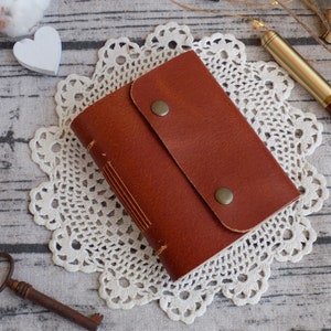 May include: A brown leather journal with a flap closure and two brass accents. The journal is sitting on a white crocheted doily.