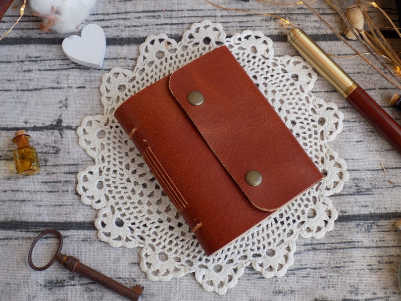 May include: A brown leather journal with a flap closure and two brass accents. The journal is sitting on a white crocheted doily.
