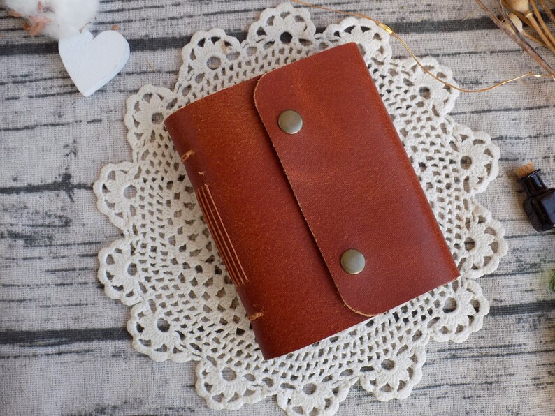 May include: A brown leather journal with a flap closure and two metal clasps. The journal is sitting on a white crocheted doily.