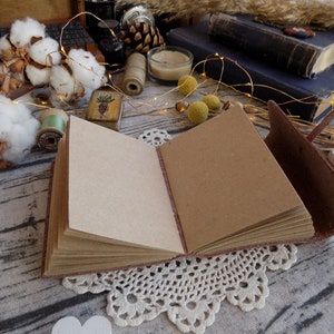 May include: A brown leather-bound journal with blank pages is open and lying on a table with a white lace doily underneath. The journal has a brown leather strap and a decorative metal clasp. The table is covered in a white fabric with a brown wooden texture.