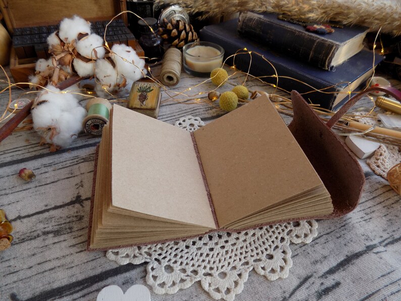 May include: A brown leather-bound journal with blank pages is open and lying on a table with a white lace doily underneath. The journal has a brown leather strap and a decorative metal clasp. The table is covered in a white fabric with a brown wooden texture.