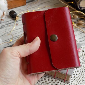 May include: A red leather-bound journal with a flap closure and a brass button. The journal is held in a hand.