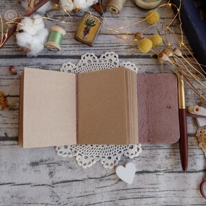 May include: A small, brown leather-bound journal with blank pages is open on a white lace doily. The journal is surrounded by various craft supplies, including cotton balls, spools of thread, and a vintage-style key.