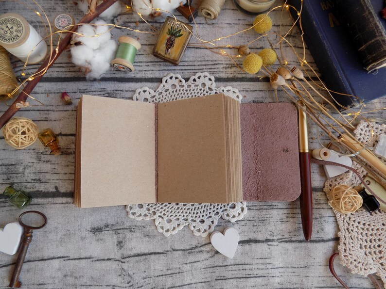 May include: A small, brown leather-bound journal with blank pages is open on a white lace doily. The journal is surrounded by various craft supplies, including cotton balls, spools of thread, and a vintage-style key.