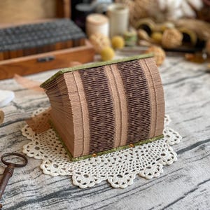 Op de afbeelding: Een gevouwen boekkunstsculptuur met afwisselende verticale banden van lichtbruine en donkerbruine papieren pagina's. Het boek is ingebonden met een groene stoffen kaft en rust op een wit gehaakt kleedje, met ingewikkelde papier-vouwtechnieken.