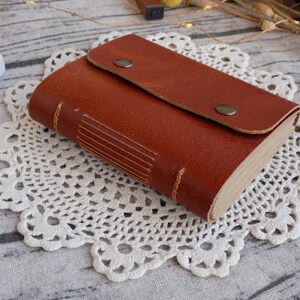 May include: A brown leather-bound journal with a flap closure and metal accents. The journal is lying on a white crocheted doily.