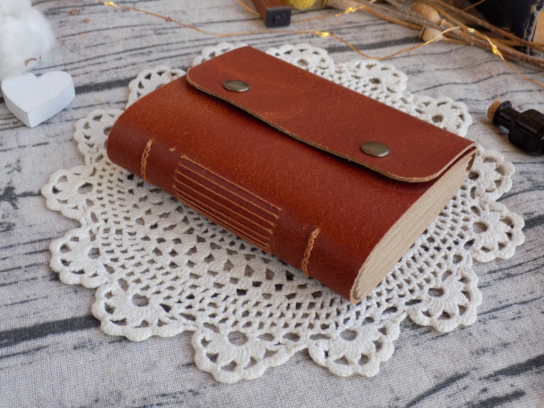 May include: A brown leather-bound journal with a flap closure and metal accents. The journal is lying on a white crocheted doily.