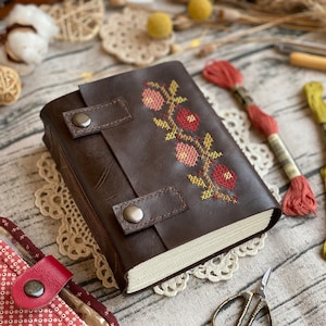 May include: A brown leather-bound journal with a floral cross-stitch design in shades of red, pink, and yellow. The journal has two leather straps with metal buckles.