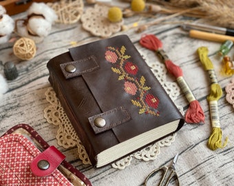 May include: A brown leather-bound journal with a floral cross-stitch design in shades of red, pink, and yellow. The journal has two leather straps with metal buckles.