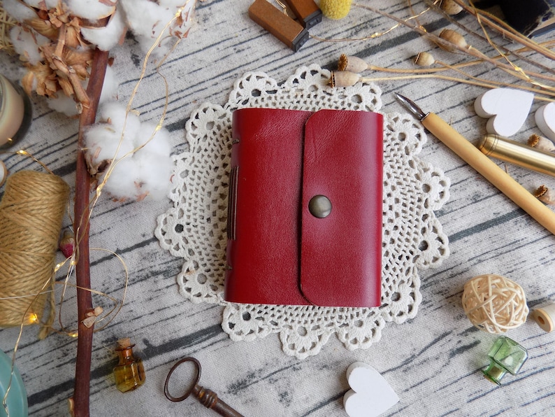 May include: A red leather journal with a flap closure and a metal button. The journal is sitting on a white doily on a wooden surface.