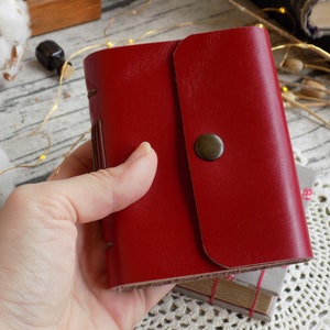 May include: A red leather-bound journal with a flap closure and a brass button. The journal is held in a hand.
