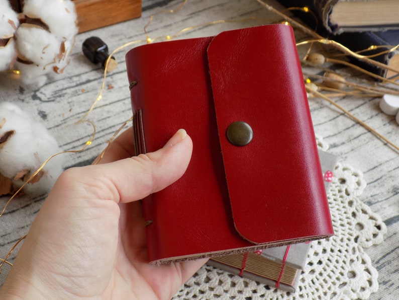 May include: A red leather-bound journal with a flap closure and a brass button. The journal is held in a hand.