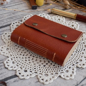 May include: A brown leather-bound journal with a flap closure and two metal snaps. The journal is sitting on a white crocheted doily.
