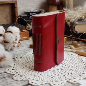 May include: A small red leather-bound journal with a decorative stitching detail on the front cover. The journal is sitting on a white crocheted doily.