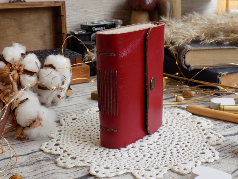 May include: A small red leather-bound journal with a decorative stitching detail on the front cover. The journal is sitting on a white crocheted doily.
