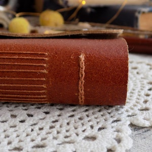 May include: Close-up of a brown leather-bound journal with visible stitching along the spine. The journal is resting on a white crocheted doily.