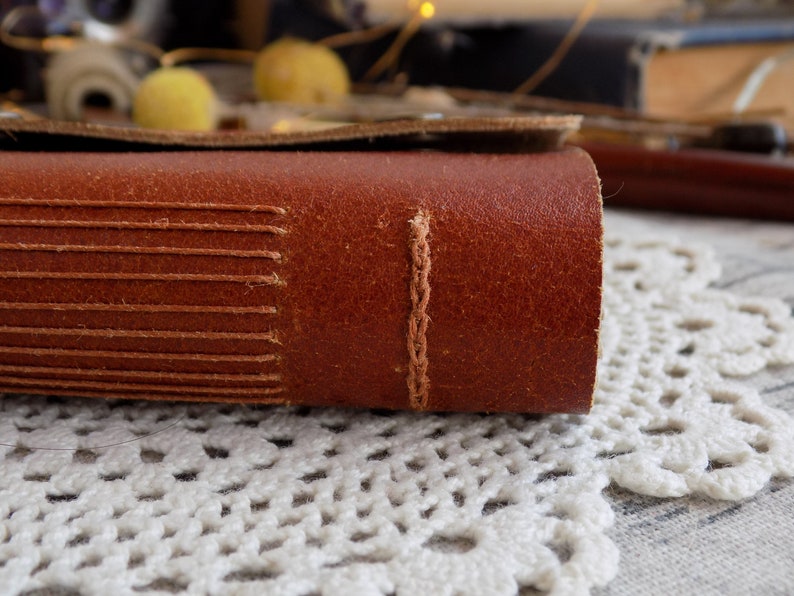 May include: Close-up of a brown leather-bound journal with visible stitching along the spine. The journal is resting on a white crocheted doily.