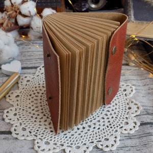 May include: A brown leather-bound journal with blank pages, open to reveal the pages. The journal is sitting on a white doily.